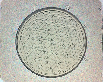May include: A silver-colored metal mold featuring a circular design of the Flower of Life pattern. The design is embossed within a circular recess, surrounded by a square frame with rounded corners and small circular rivets.