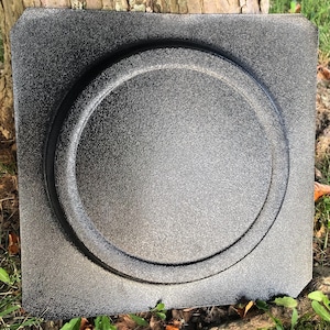 May include: A square, black, textured concrete stepping stone with a circular recessed design in the center. The stone has a rough, speckled surface and is likely used for outdoor landscaping or garden pathways.