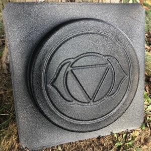 May include: A black and gray textured square with a raised circular design. The design features a stylized eye with a triangle inside.