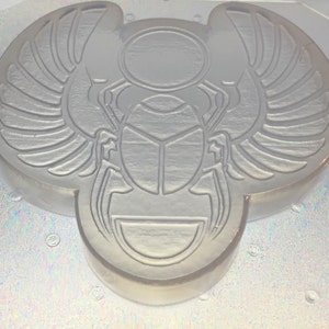 May include: A clear, resin mold in the shape of a scarab beetle with outstretched wings. The design features detailed lines and a circular sun emblem above the beetle. The mold is likely for creating decorative items or jewelry.
