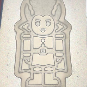 Puede incluir: Un molde de plástico de color claro con forma de personaje de dibujos animados. El personaje tiene una cara redonda con ojos grandes, una pequeña sonrisa y un cuello puntiagudo. El molde está diseñado con líneas detalladas.