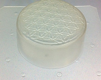 May include: A clear, cylindrical object with a geometric pattern on the top surface. The object is made of a translucent material and sits on a textured, light-colored surface. The top surface features a repeating design.