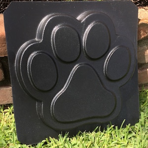 May include: Black plastic mold for making concrete paw prints. The mold is shaped like a dog paw with four toes and a pad.