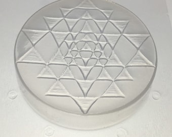 May include: A white, round, decorative object with a raised, intricate geometric design. The design is a series of overlapping triangles that create a star-like pattern.