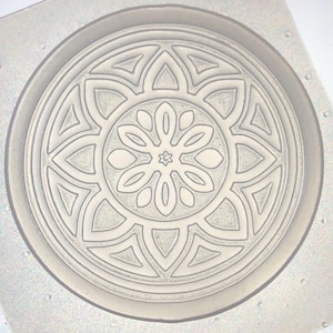 May include: A circular, light gray mold with a detailed floral design. The design features a central starburst surrounded by petal-like shapes and geometric patterns. The mold is set within a square frame.