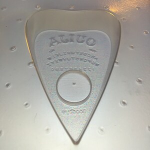 May include: A clear, heart-shaped Ouija planchette with the word "Ouija" at the top. The planchette has a circular opening and is surrounded by letters and numbers. The planchette is made of a clear, iridescent material.
