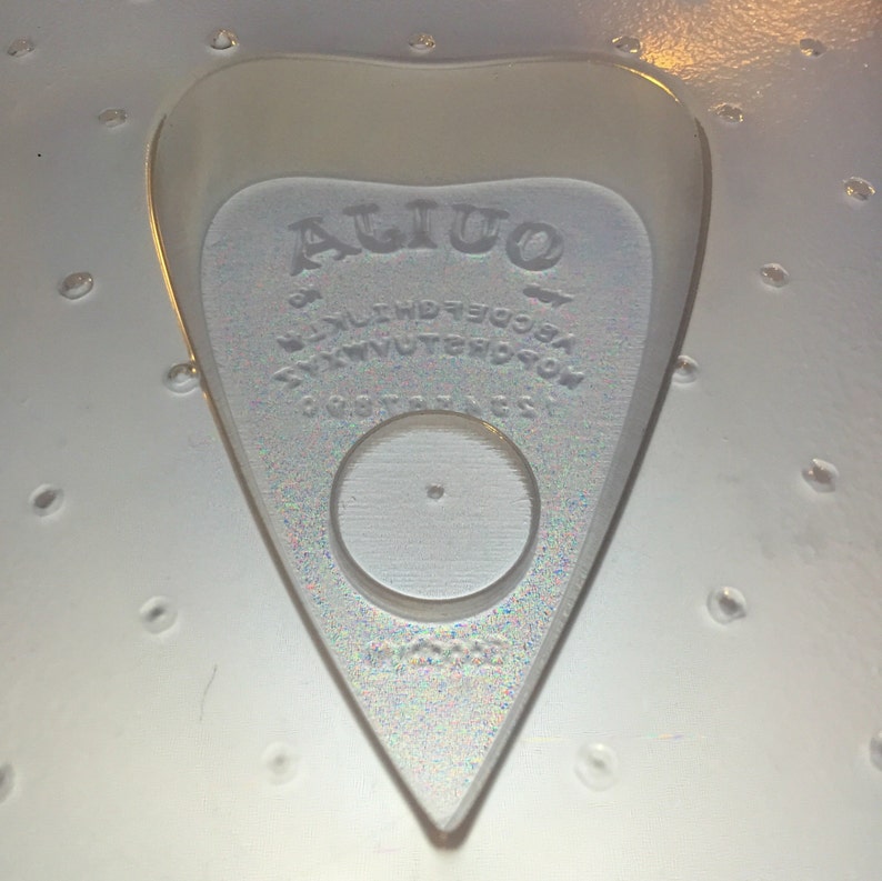May include: A clear, heart-shaped Ouija planchette with the word "Ouija" at the top. The planchette has a circular opening and is surrounded by letters and numbers. The planchette is made of a clear, iridescent material.