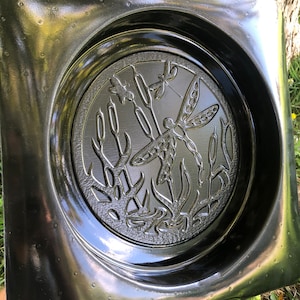 May include: A black square concrete mold with a raised design of a dragonfly and reeds. The mold is designed to create a decorative concrete piece for a garden.