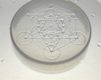 May include: A clear, round, glass coaster with a white Metatron's Cube design. The geometric pattern is raised and detailed, set against a translucent background. The coaster is on a white surface.