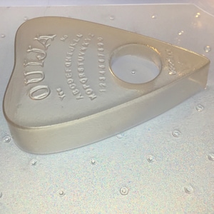 May include: A translucent, heart-shaped Ouija planchette. The planchette features the word "Ouija" at the top, with the alphabet, numbers, and the words "yes" and "no" embossed on the surface. It has a circular opening for use.