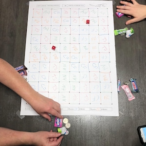 Könnte beinhalten: Ein Brettspiel mit einem Raster aus Quadraten, die jeweils handschriftliche Buchstaben und Zahlen enthalten, auf einer dunklen Oberfläche befestigt. Zwei rote Würfel liegen auf dem Brett. Hände halten Süßigkeiten und Münzen. Buntstifte sind sichtbar.