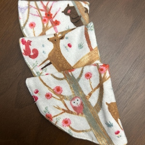 May include: Three white fabric items with a woodland animal print. The print features brown deer, squirrels, and owls among flowering branches. The items are arranged in a layered stack, showcasing the repeating pattern.