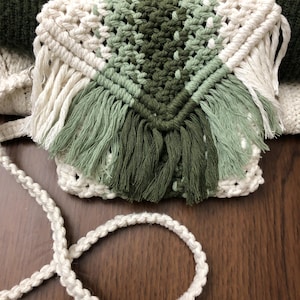 May include: A handmade macrame crossbody bag in cream, and shades of green. The bag features a woven design with a fringe detail and a cream-colored rope strap. The bag is a stylish accessory for carrying essentials.