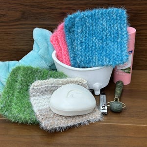 Puede incluir: Una colección de productos para el baño y el cuerpo, que incluye un jabón blanco, cortaúñas, un rodillo facial y varios paños de lavado coloridos. Los paños de lavado son en tonos de azul, rosa, verde y tonos neutros. Un pequeño recipiente blanco en forma de bañera contiene algunos de los paños.