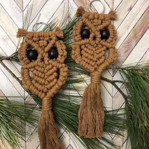 May include: Two brown macrame owl keychains with black bead eyes and a tassel. The keychains are hanging on a branch with green pine needles, set against a light brown and white chevron pattern background.