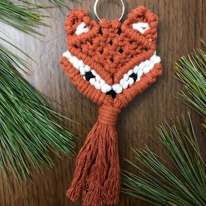 May include: A handmade macrame fox keychain in shades of orange, white, and black. The fox has a tassel tail and a silver key ring. The keychain is displayed on a wooden surface with green pine needles.