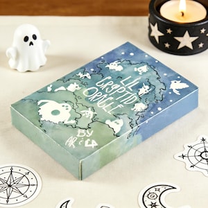 Könnte beinhalten: Rechteckige Schachtel für das "Lil Cryptid Oracle" Kartenspiel, mit einem Aquarelldesign von skurrilen Kreaturen und dem Text "Lil Cryptid Oracle" in Weiß. Die Schachtel ist in Blau- und Grüntönen gehalten, mit einer weißen Geisterfigur und einem Kerzenhalter im Hintergrund.