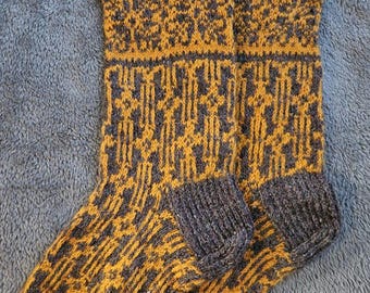 May include: A pair of knitted socks with a geometric pattern in shades of gold and dark gray. The socks feature a ribbed cuff and a patterned design that covers the entire sock. The socks are made of a soft, warm material.