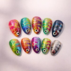 May include: A collection of ten colorful, fish-themed nail art designs. Each nail features a unique design with vibrant hues, glitter accents, and a fish eye. The nails are arranged on a white surface.