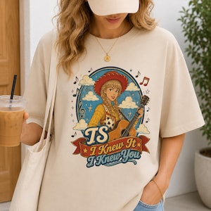 May include: A beige short-sleeve t-shirt featuring a graphic of a blonde person in a red cowboy hat playing an acoustic guitar. The design includes clouds, musical notes, and the text "TS I Knew It I Knew You" on a red banner.