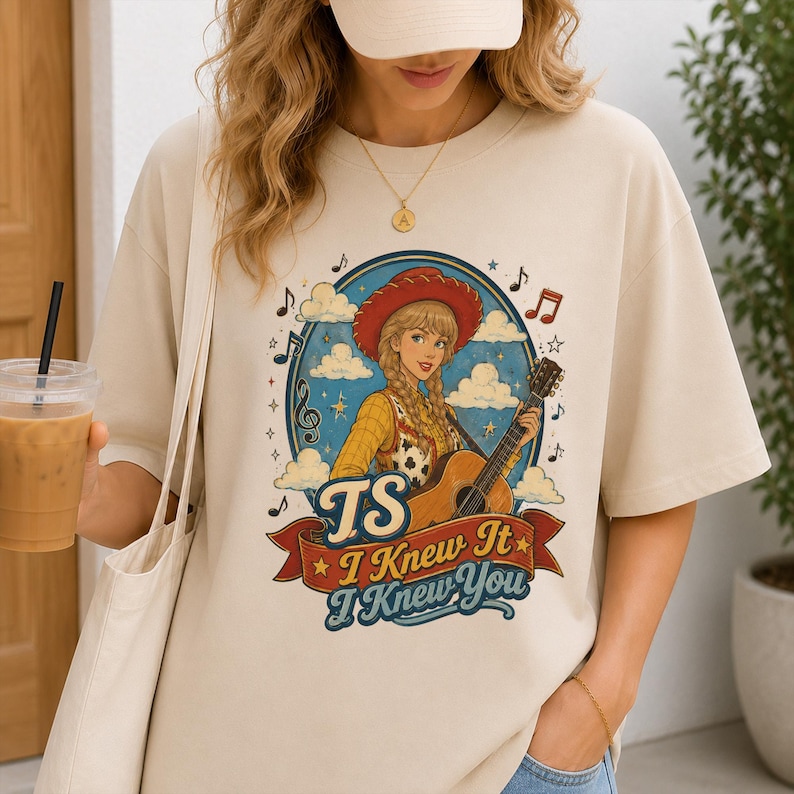 May include: A beige short-sleeve t-shirt featuring a graphic of a blonde person in a red cowboy hat playing an acoustic guitar. The design includes clouds, musical notes, and the text "TS I Knew It I Knew You" on a red banner.