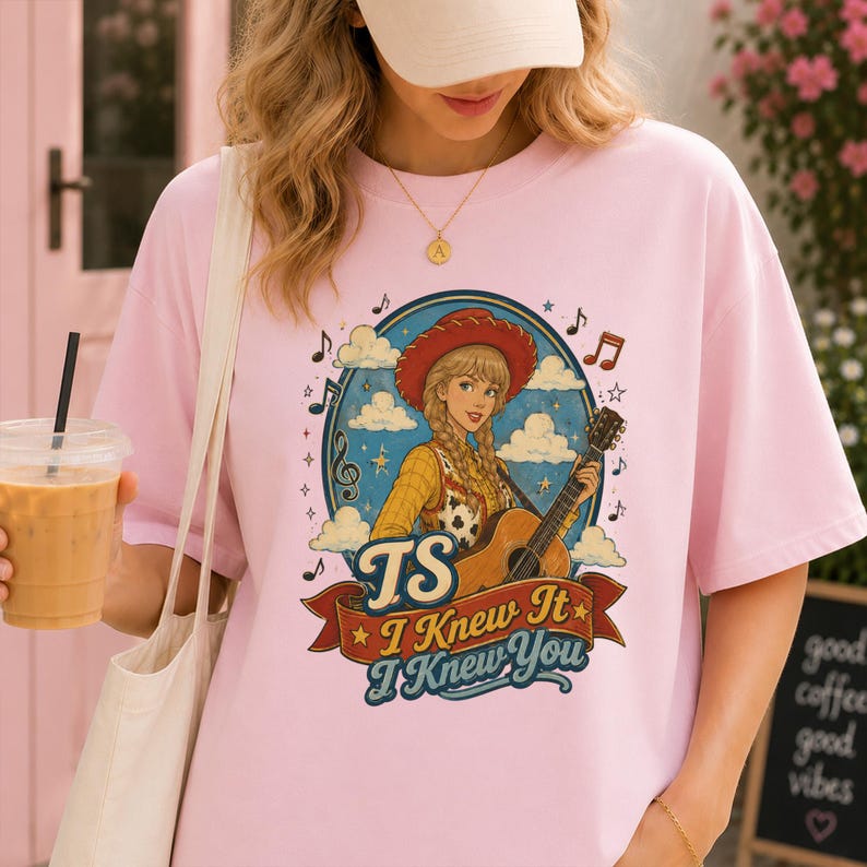 May include: A light pink t-shirt featuring a graphic illustration of a person with blonde braids wearing a red cowboy hat and yellow shirt, playing an acoustic guitar. The text reads TS I Knew It I Knew You, surrounded by musical notes and clouds.