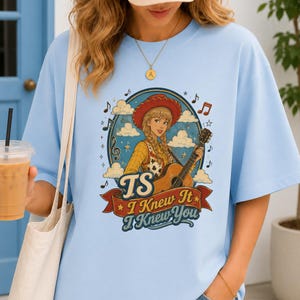 May include: A light blue t-shirt featuring a graphic of a cowgirl with blonde braids playing an acoustic guitar inside a circular frame with clouds and musical notes. Below the illustration, a red banner displays the text TS I Knew It I Knew You.