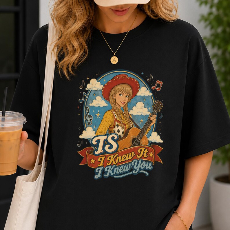 May include: A black t-shirt featuring a graphic of a blonde person in a cowgirl hat and yellow shirt playing an acoustic guitar. The design includes clouds, musical notes, and the text "TS I Knew It I Knew You" on a red banner.