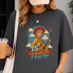 May include: A charcoal gray t-shirt featuring a graphic of a blonde person in a red cowboy hat playing an acoustic guitar. The design includes clouds, musical notes, and the text TS I Knew It I Knew You on a red banner at the bottom.