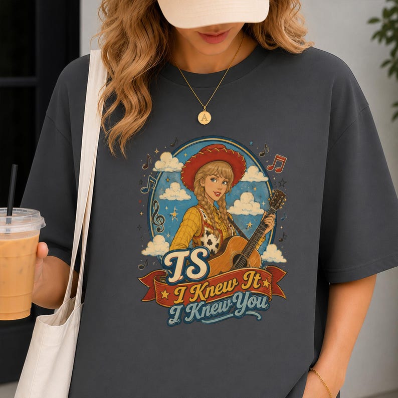 May include: A charcoal gray t-shirt featuring a graphic of a blonde person in a red cowboy hat playing an acoustic guitar. The design includes clouds, musical notes, and the text TS I Knew It I Knew You on a red banner at the bottom.