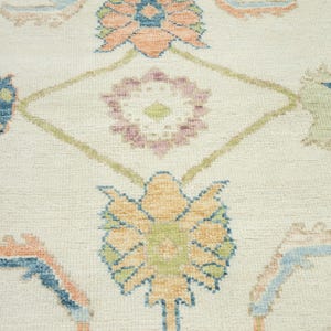 May include: Close-up of a cream-colored rug with a floral pattern. The design features stylized flowers in shades of orange, blue, green, and purple, set against a cream background. The rug has a textured appearance.
