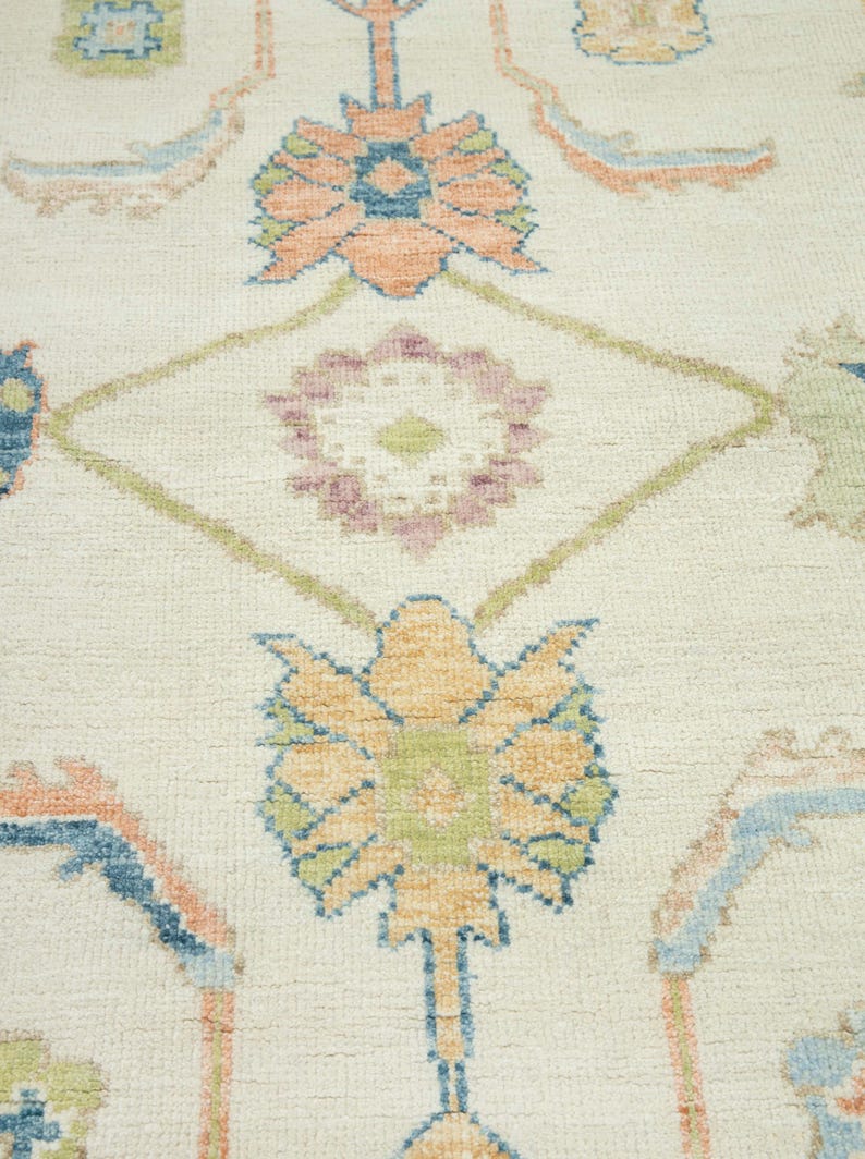 May include: Close-up of a cream-colored rug with a floral pattern. The design features stylized flowers in shades of orange, blue, green, and purple, set against a cream background. The rug has a textured appearance.