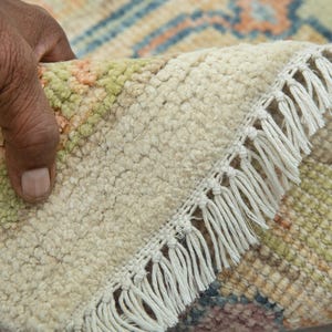 May include: Close-up of a hand lifting a corner of a beige rug with a cream fringe. The rug has a textured surface with a pattern of green, orange, and blue geometric shapes. The rug's edge is visible.