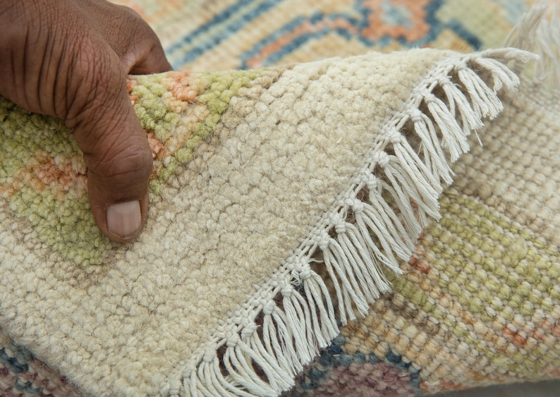 May include: Close-up of a hand lifting a corner of a beige rug with a cream fringe. The rug has a textured surface with a pattern of green, orange, and blue geometric shapes. The rug's edge is visible.