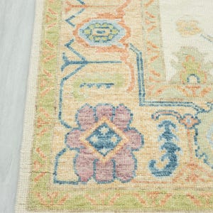 May include: Close-up of a beige area rug with a detailed floral pattern. The rug features a border with green, blue, and peach accents. The central design includes a large, multi-colored floral motif with a diamond center. The rug has a fringe.