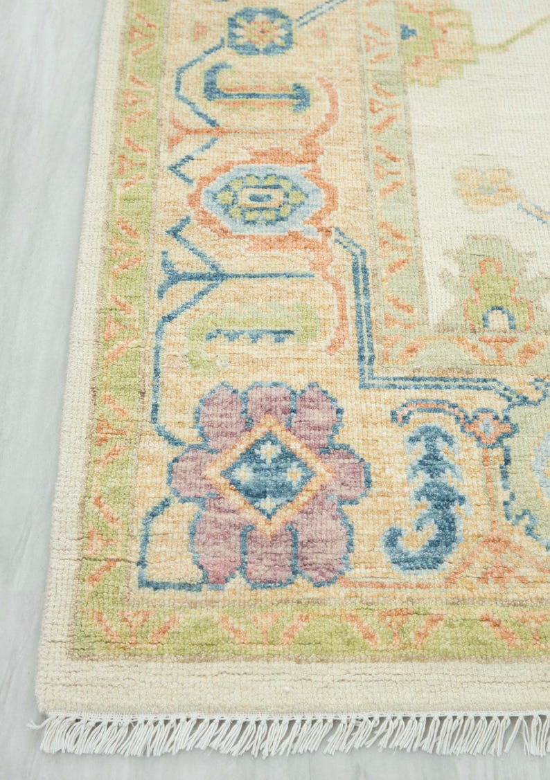 May include: Close-up of a beige area rug with a detailed floral pattern. The rug features a border with green, blue, and peach accents. The central design includes a large, multi-colored floral motif with a diamond center. The rug has a fringe.