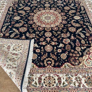 May include: A rectangular area rug featuring a deep navy blue central field with an intricate cream, red, and sage green floral medallion and vine pattern. The rug has a wide cream border with floral motifs and white fringe along the edges.