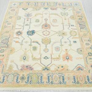 May include: Cream-colored area rug with a detailed floral pattern in shades of blue, peach, and green. The rug has a border with repeating floral and geometric designs. The rug is rectangular and has a fringe along the edges.