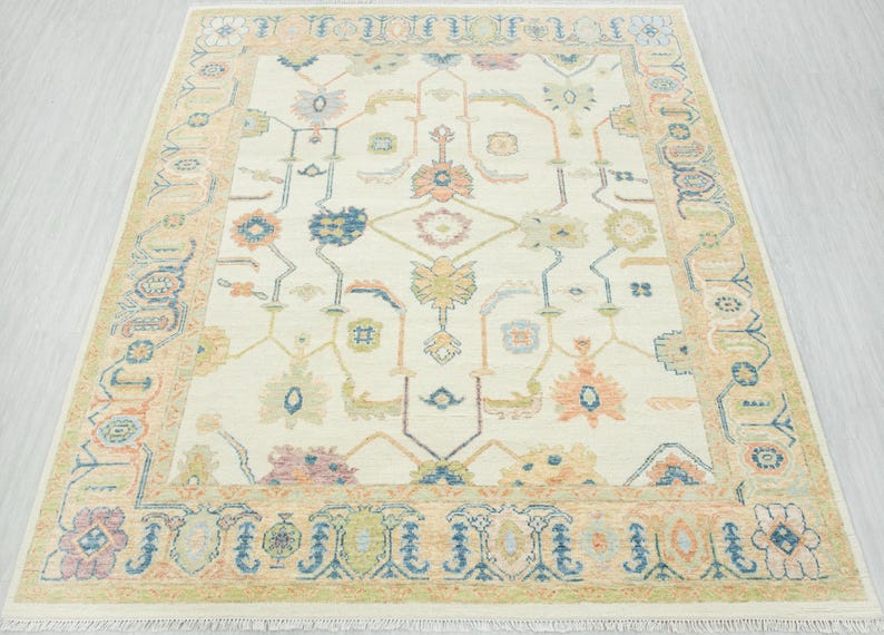 May include: Cream-colored area rug with a detailed floral pattern in shades of blue, peach, and green. The rug has a border with repeating floral and geometric designs. The rug is rectangular and has a fringe along the edges.