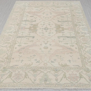 May include: Cream-colored area rug with a detailed floral and geometric pattern. The rug features a central design with symmetrical elements in soft pink and green, framed by a border with similar motifs. The rug is rectangular and appears to be made of woven material.