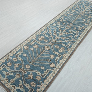 Peut inclure: Un long tapis rectangulaire de couloir présentant un fond bleu profond avec un motif répétitif d'arbres stylisés et d'éléments floraux géométriques beiges et bruns. Le tapis possède une bordure décorative et des franges blanches aux extrémités.