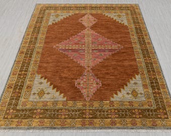 May include: A rectangular rug with a central diamond design in shades of pink and red, set against a rust-colored background. The border features a repeating floral pattern in gold and brown, with a light gray and yellow geometric design.