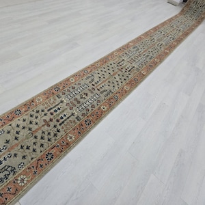 Puede incluir: Una alfombra larga y estrecha con un campo central beige y patrones geométricos intrincados. La alfombra presenta un borde en tonos naranja y marrón, con motivos florales y geométricos. El diseño de la alfombra es simétrico.