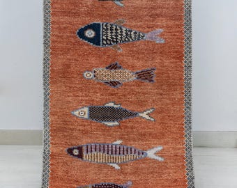 May include: A rectangular runner rug with a burnt orange background featuring eight horizontal rows of stylized fish in various colors including blue, brown, and purple. The rug has a gray and white patterned border and fringed ends.