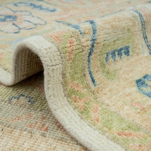 May include: Close-up of a rolled-up area rug with a cream-colored border. The rug features a floral pattern in shades of blue, green, and peach on a beige background. The texture of the rug is visible, highlighting the woven design.
