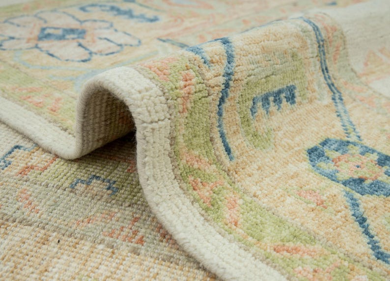 May include: Close-up of a rolled-up area rug with a cream-colored border. The rug features a floral pattern in shades of blue, green, and peach on a beige background. The texture of the rug is visible, highlighting the woven design.