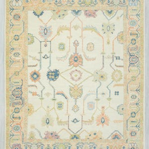 May include: Cream-colored area rug with a detailed floral and geometric pattern. The rug features a central design with symmetrical floral motifs in shades of blue, peach, and green, framed by a border in a similar color palette. The rug has a fringed edge.