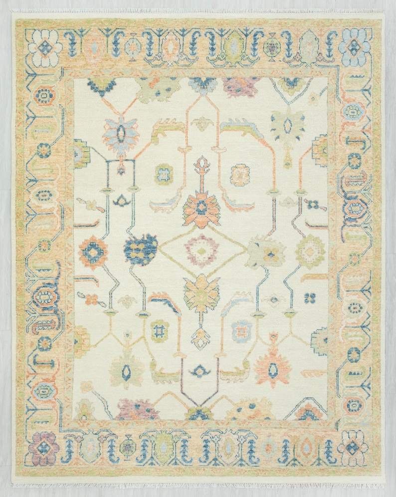 May include: Cream-colored area rug with a detailed floral and geometric pattern. The rug features a central design with symmetrical floral motifs in shades of blue, peach, and green, framed by a border in a similar color palette. The rug has a fringed edge.