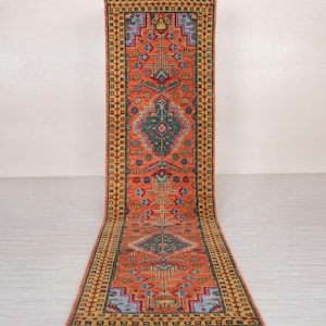 May include: An orange runner rug with a geometric pattern. The rug features a central diamond design in teal, surrounded by red, blue, and yellow accents. The border is a repeating pattern of yellow and blue squares. The rug is approximately 8 feet long.