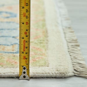 May include: Close-up of a section of a rug with a cream-colored border and fringe. The rug has a light blue and peach pattern. A yellow measuring tape shows the rug is approximately 13 inches long.